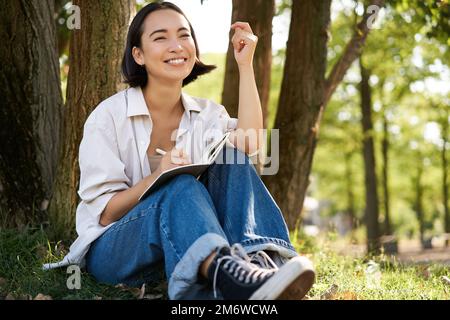 Porträt einer jungen asiatischen Frau, die in ihr Notizbuch schreibt, ihre Gedanken auf Papier in Tagebüchern ausdrückt, lächelt und unter dem Baum sitzt Stockfoto