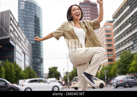 Porträt einer tanzenden asiatischen Frau, die die Hände hochhebt und sich glücklich fühlt, in der Stadt posiert, triumphiert, Freude fühlt Stockfoto