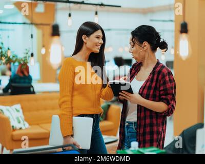 Porträt von zwei Geschäftsfrauen, die miteinander sprechen, während sie in einem modernen Büro mit ihren Kollegen mit Lapto stehen Stockfoto