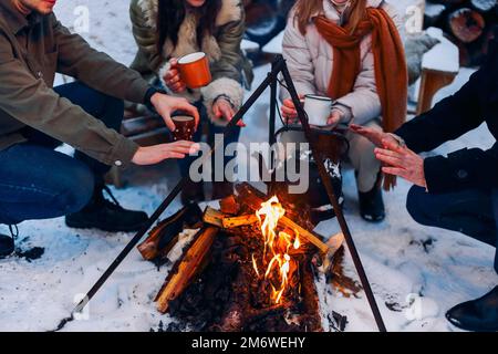 Eine Gruppe von Freunden versammelt sich am Lagerfeuer im Garten, trinkt Tee und wärmt sich die Hände Stockfoto