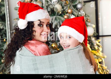 Mutter und Kind tragen weihnachtsmannmützen, die sich unter einer Strickdecke auf dem Boden neben dem Weihnachtsbaum umarmen Stockfoto
