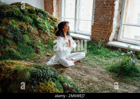 Großes Foto einer optimistischen brünetten Dame, die auf dem Rasen sitzt. Rest tragen Sie weiße bequeme Kleidung, Oberteil und Hose, innen Stockfoto