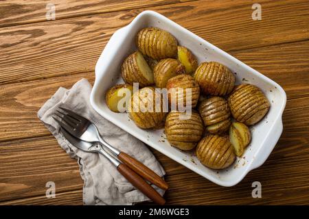 Hasselback gebackene Kartoffeln in weißer Keramik-Auflaufform auf rustikalem Holztisch Hintergrund Stockfoto