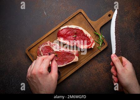 Männliche Hand hält Messer über zwei rohen Fleisch Rindfleisch Steaks Draufsicht Stockfoto