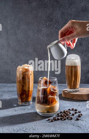 Milch wird auf einem dunklen Tisch in Eiskaffee gegossen. Eiskaffee in einem großen Glas mit Sahne und Kaffeebohnen. Kalter Cof Stockfoto