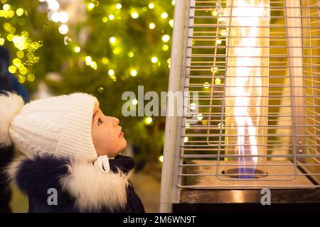 Ein kleines Mädchen mit warmem Hut sieht die Flamme eines Gasbrenners, der sich auf einem Weihnachtsmarkt in der Stadt erwärmt. Lichter der Girlanden rein Stockfoto