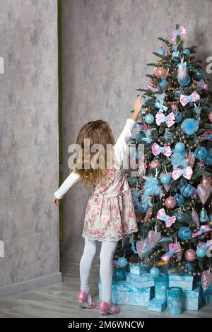 Ein kleines Mädchen greift mit der Hand nach einem Weihnachtsbaumspielzeug. Weihnachtsdekor, wartet auf einen Urlaub und ein Wunder. Neues Jahr. Wh Stockfoto