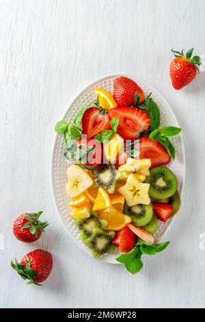 Bunte reife Früchte und Beeren auf dem Teller, Draufsicht auf weißem Hintergrund. Vegetarisches Speisenkonzept. Erdbeere, Kiwi, Apfel und Stockfoto