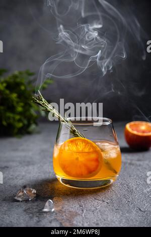 Negroni mit rauchendem Rosmarin. Negroni mit Eis und Orange auf dunklem Hintergrund. Negroni-Cocktail in einem Glas mit rauchendem Rosema Stockfoto