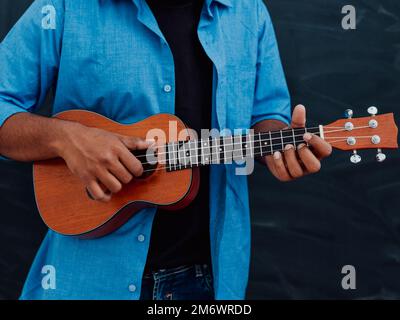 Indischer junger Mann in einem blauen Hemd und einer Brille, der vor der Schultafel Gitarre spielt Stockfoto