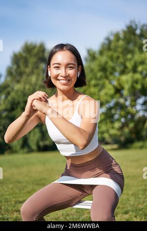 Asiatische Fitness-Mädchen tut Kniebeugen im Park, mit Widerstand Band, Stretching Yoga-Seil für das Training an der frischen Luft Stockfoto