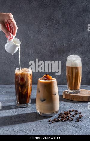 Milch wird auf einem dunklen Tisch in Eiskaffee gegossen. Eiskaffee in einem großen Glas mit Sahne und Kaffeebohnen. Kalter Cof Stockfoto