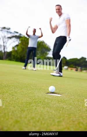 Der perfekte Putt. Ein kleiner Winkel, bei dem sich ein Golfball dem Loch nähert, während zwei Golfer zuschauen. Stockfoto