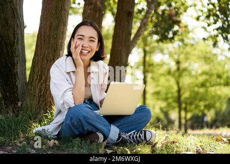 Porträt einer jungen asiatischen Frau, die im Park neben einem Baum sitzt, an einem Laptop arbeitet und draußen Computer benutzt Stockfoto