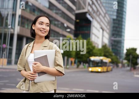 Schöne asiatische Mädchen lächelt als pendelt zur Arbeit, steht auf der Straße mit Laptop und Notebook Stockfoto