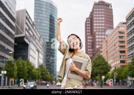 Portrait einer glücklichen asiatischen Frau steht mit einem Tablet in der Nähe der Straße, jubelt, hebt die Hand hoch in Triumph, feiert Stockfoto