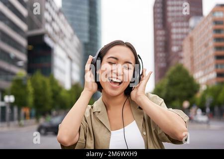 Tanzendes Mädchen glücklich in der Stadt fühlen. Asiatische Frau tanzt und hört Musik in Kopfhörern, posiert auf der Straße Stockfoto