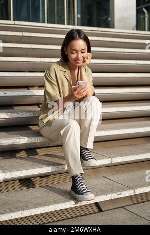 Vertikale Aufnahme einer asiatischen Frau, eine Studentin sitzt auf einer Treppe in der Stadt, schaut auf den Bildschirm des Mobiltelefons und lächelt mit der Smartphone-App Stockfoto