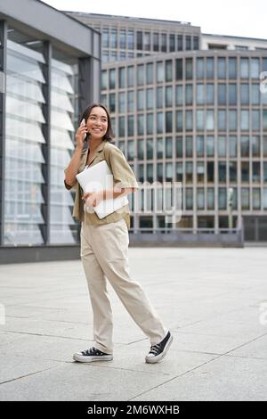 Vertikales Porträt einer stilvollen asiatischen Schülerin, die beim Gehen mit dem Handy spricht und ein Notebook in der Hand hält Stockfoto