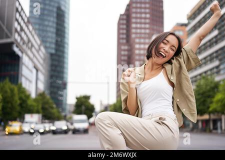 Porträt einer glücklichen asiatischen Frau, tanzen und Freude fühlen, triumphieren, die Hand in Siegesgeste hochheben, auf der Straße feiern Stockfoto