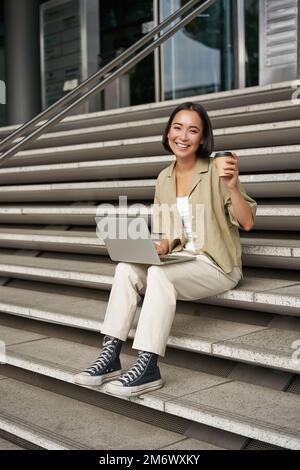 Vertikale Aufnahme einer lächelnden Studentin, asiatische Frau sitzt auf der Treppe des Universitätscampus und trinkt Kaffee, macht ihre Hausaufgaben an mir Stockfoto