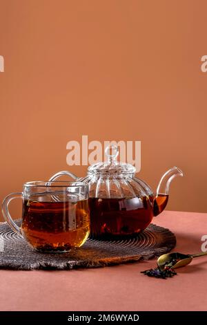 Glasteekanne, Tasse Tee und Teeblätter auf braunem Hintergrund. Heißer Kräutertee ist in der Glasteekanne auf dem Tisch. Speicherplatz kopieren. Stockfoto