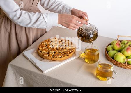 Eine Frau, die Kräutertee einschenkt. Hausgemachter Apfelkuchen auf einem Schneidebrett auf einem Familientisch. Herbstbacken Stockfoto