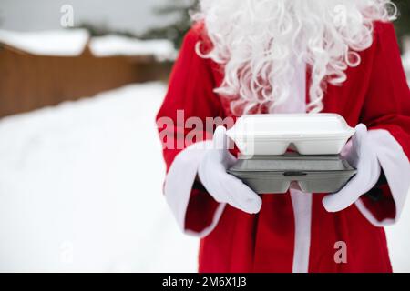 Lebensmittelliefercontainer in den Händen des Weihnachtsmanns im Freien im Schnee. Weihnachtsabend-Angebot. Fertig zubereitete heiße Bestellung, Disposa Stockfoto