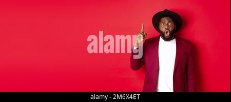 Mode- und Partykonzept. Aufgeregter Schwarzer, der eine Idee hat, den Finger rührt, um einen Vorschlag zu sagen, in stilvollem Hut und Blazer steht Stockfoto