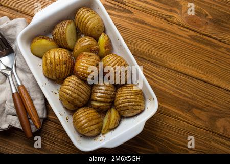 Hasselback gebackene Kartoffeln in weißer Keramik-Auflaufform auf rustikalem Holztisch Hintergrund Stockfoto