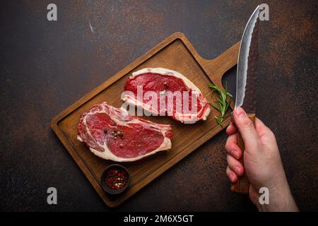 Männliche Hand hält Messer über zwei rohen Fleisch Rindfleisch Steaks Draufsicht Stockfoto