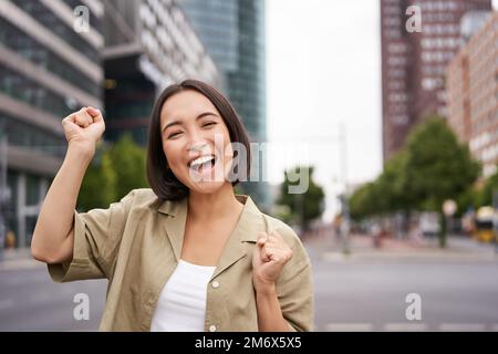 Glückliches asiatisches Mädchen gewinnt und freut sich, feiert auf den Straßen der Stadt, tanzt vor Freude, triumphiert und lächelt Stockfoto