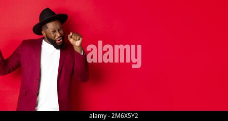 Weihnachten und Menschen Konzept. Happy Black Mann feiert Neujahr und tanzen, trägt Party-Outfit, roten Hintergrund Stockfoto