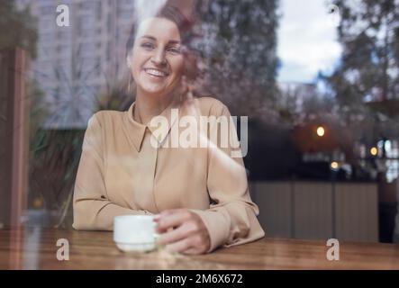 Ehrliches Porträt einer lachenden jungen Frau, die lässige Kleidung trägt und eine Tasse Kaffee hält Stockfoto