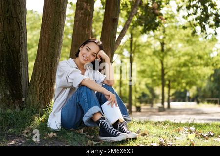 Frau sitzt im Park mit ihrem Lieblingsbuch, lehnt sich an sonnigen Tagen auf einen Baum im Schatten, genießt die Natur und das ruhige, entspannende atmos Stockfoto