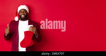 Schöner bärtiger Kerl erhalten Weihnachtsgeschenk in Socke, zeigt Daumen-up und lächelnd, stehen vor rotem Hintergrund Stockfoto