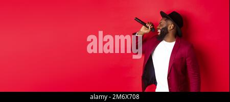 Leidenschaftlicher schwarzer Sänger vor rotem Hintergrund, singt ins Mikrofon, trägt Party-Outfit, steht über rot Stockfoto
