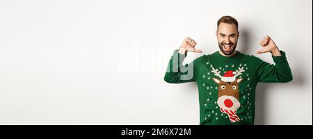 Winterferien und weihnachten. Selbstbewusster junger Mann, der enthusiastisch aussieht, stolz auf sich zeigt, über weißem Backgr steht Stockfoto