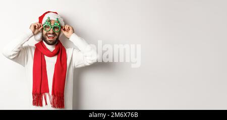 Konzept für Weihnachten, Neujahr und Feierlichkeiten. Glücklicher Mann, der lacht, Weihnachtsmannmütze und Partybrille trägt, über dem weißen Backgr steht Stockfoto