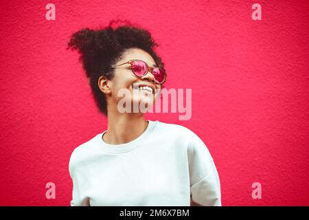 Junge Frau aus afrikanischer Ethnie in stilvoller Sonnenbrille, mit lockigen Haaren, die in einem hohen Pferdeschwanz zusammengebunden sind, und mit breitem Lächeln wegschaut Stockfoto