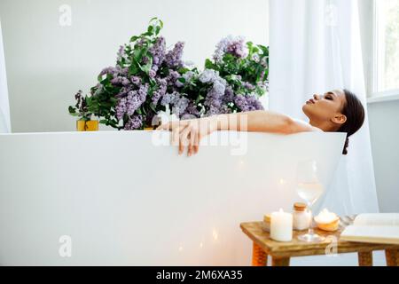 Junge brünette Frau liest das Buch und trinkt Weißwein im Bad mit Schaum und Kerzen Stockfoto