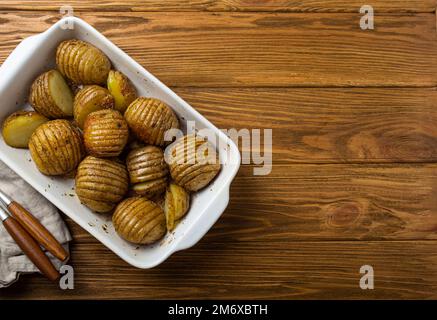 Hasselback gebackene Kartoffeln in weißer Keramik-Auflaufform auf rustikalem Holztisch Hintergrund Stockfoto