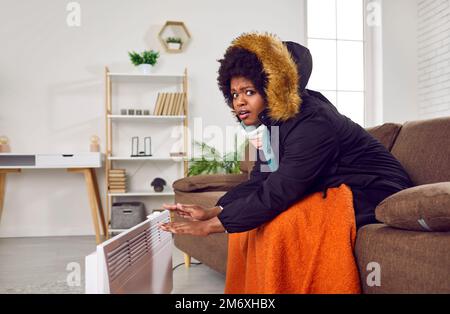Eine afroamerikanische Frau im Wintermantel auf dem Sofa und wärmt sich die Hände mit einem elektrischen Heizgerät Stockfoto