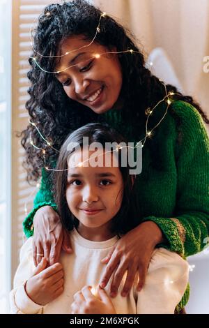 Brünett Mädchen und lange lockige afroamerikanische Mutter Pose mit leuchtenden Girlande auf dem Kopf und lächeln glücklich gegen Weihnachtsbaum Stockfoto