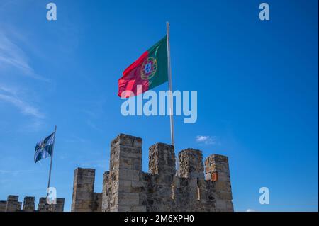 Castelo de Sao Jorge alias St. George Castle mit dem Burggraben, den Wachtürmen, den Zinnen, den Festungsmauern und der winkenden portugiesischen Flagge, die in Lissabon, Portugal, gefangen genommen wurde. Stockfoto