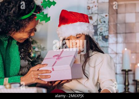 Brünette langhaarige Tochter in Santa Hut öffnet festliche Box mit Geschenken sitzen in der Nähe afroamerikanische Mutter gegen Weihnachtsbaum Stockfoto