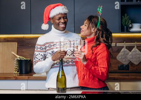 Eine junge weiße Frau mit lockigem Haar und ein schwarzer Freund, der einen festlichen Pullover trägt, feiern Weihnachten, indem sie Champagner aus Gläsern in der Küche trinkt Stockfoto