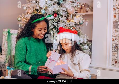 Lockige afroamerikanische Frau und brünette langhaarige Tochter tragen Urlaubshüte offene Geschenkbox sitzen gegen Weihnachtsbaum Stockfoto