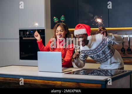 Ein glückliches junges Paar sieht sich festliche Shows mit dem Laptop an und feiert Weihnachten mit Glitzer und Champagner in der Küche Stockfoto