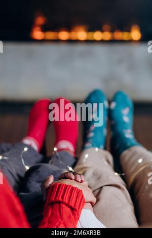 Ein paar afroamerikanische Männer und Frauen mit gemischten Rassen halten sich die Hände in festlichen, bunten Socken, die auf dem Sofa neben dem Kamin sitzen Stockfoto